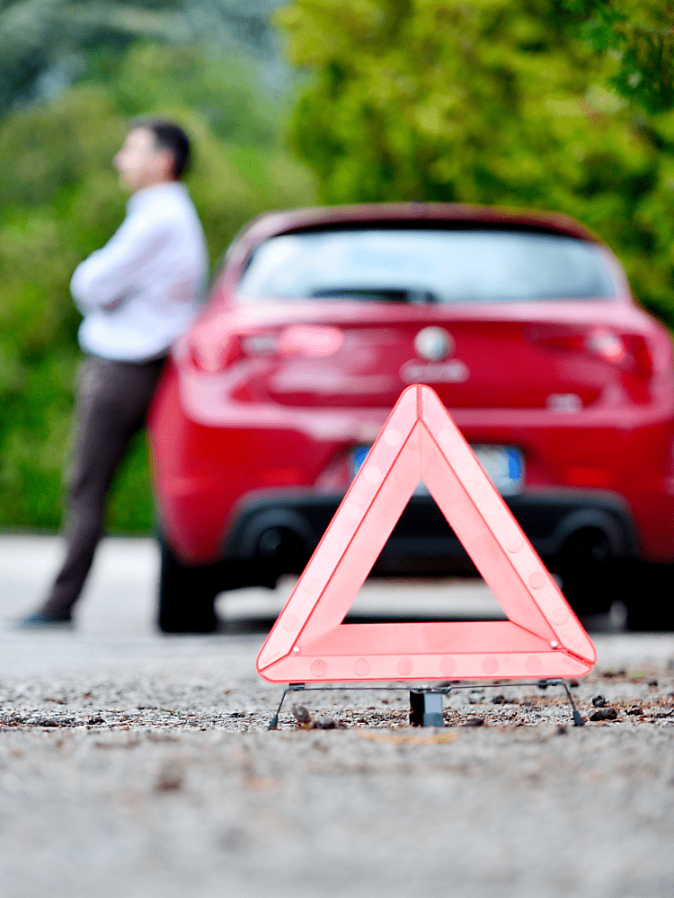 Red car broken down on roadside, man leaning against it; warning triangle in foreground.