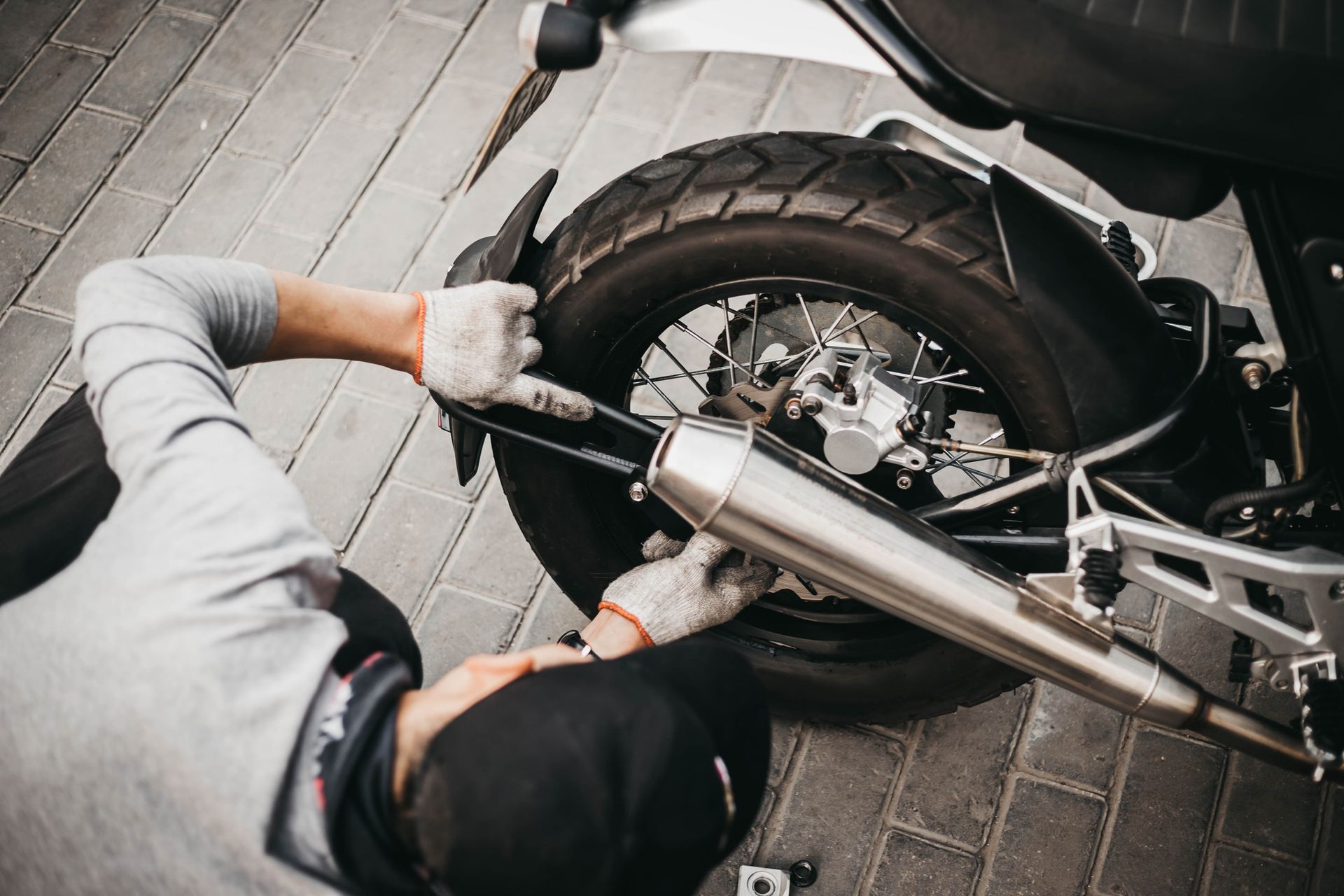 Person in gray shirt and gloves working on motorcycle exhaust on a brick surface.