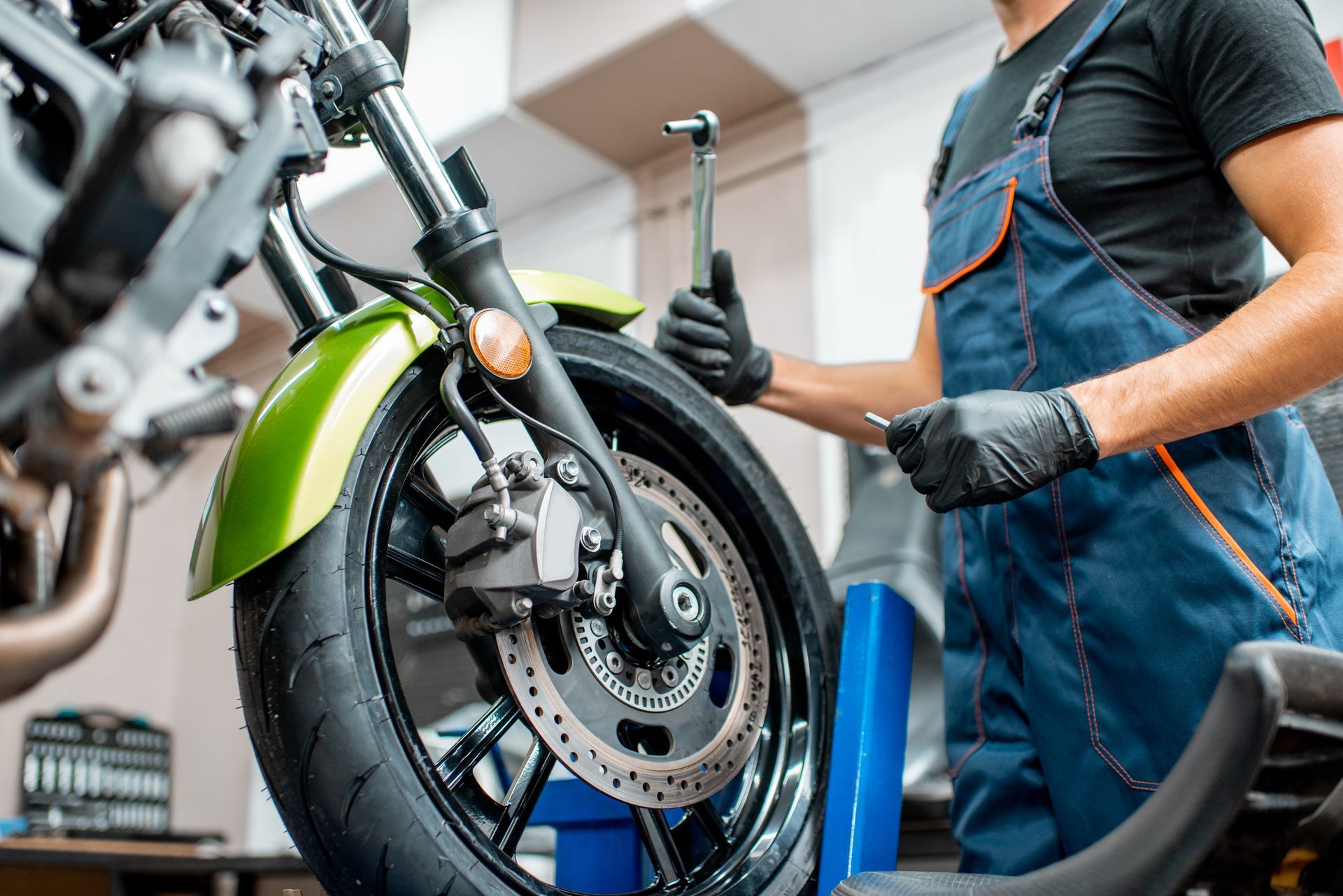 Mechanic in overalls working on a green motorcycle wheel with tools in hand.