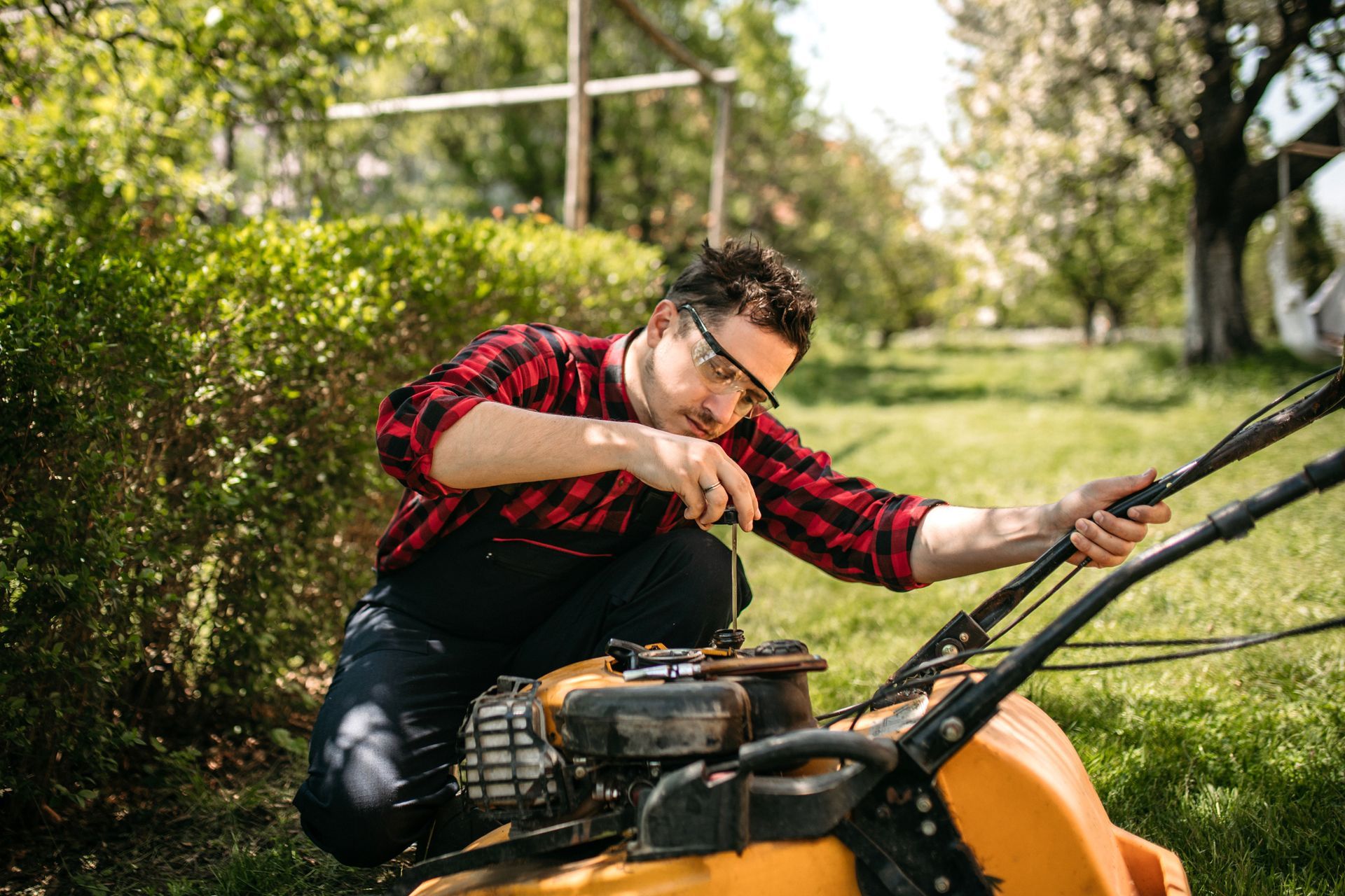 Man in plaid shirt repairs a yellow lawnmower in a grassy yard.