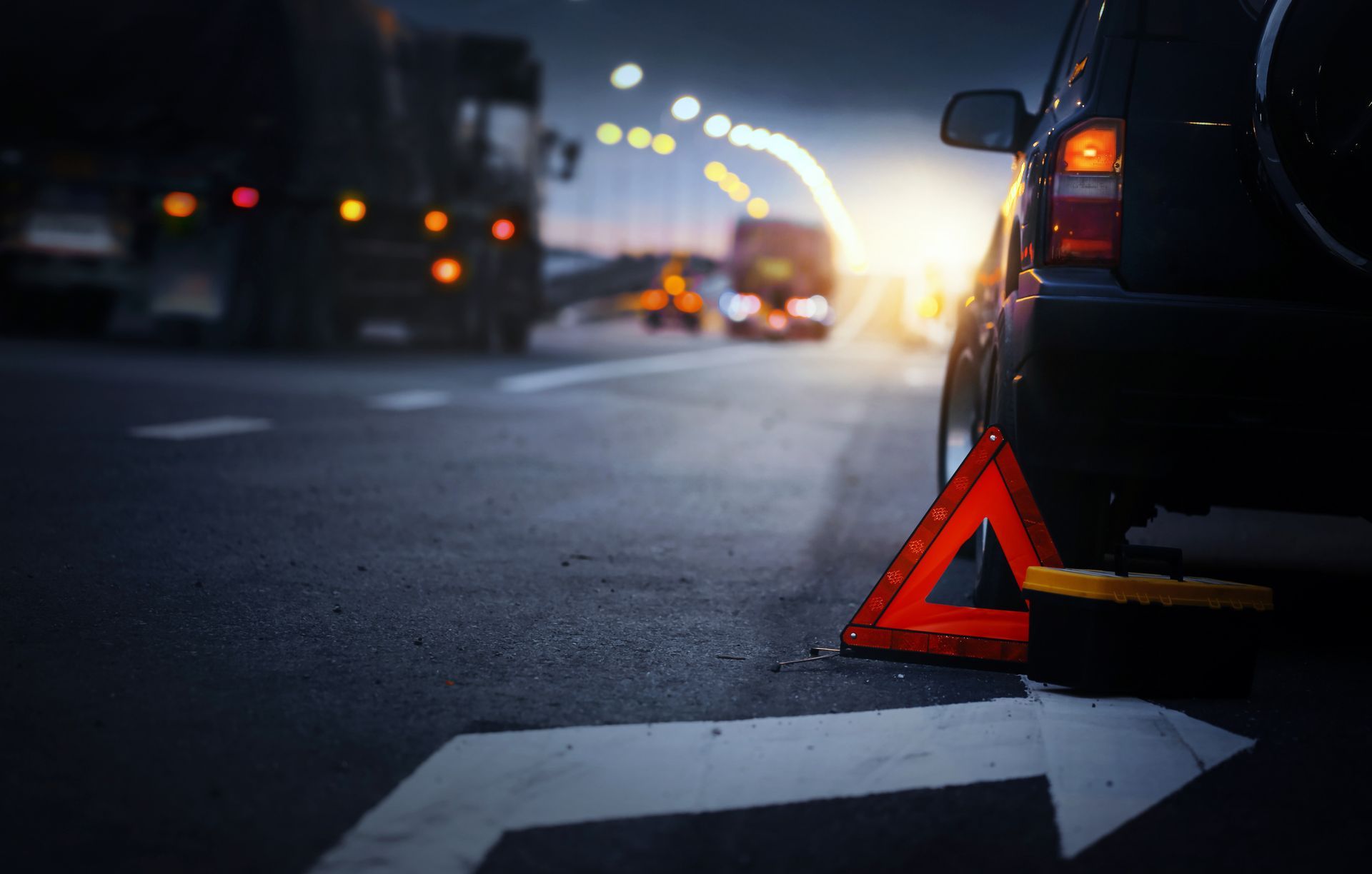Car with hazard triangle on road at night, headlights, truck in background.
