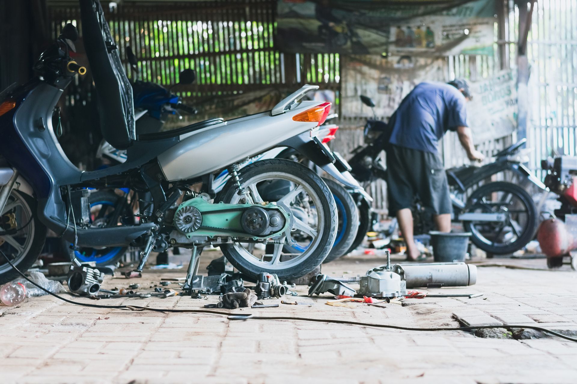 Motorcycle mechanic working on a scooter in a cluttered garage, with spare parts and tools on the floor.