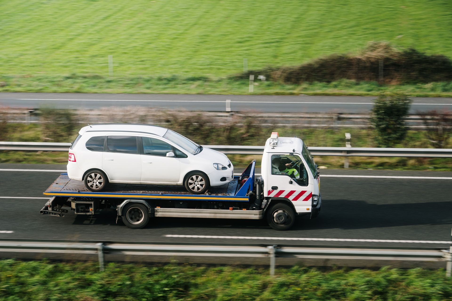 White car on a flatbed tow truck traveling on a highway, with green fields in the background.