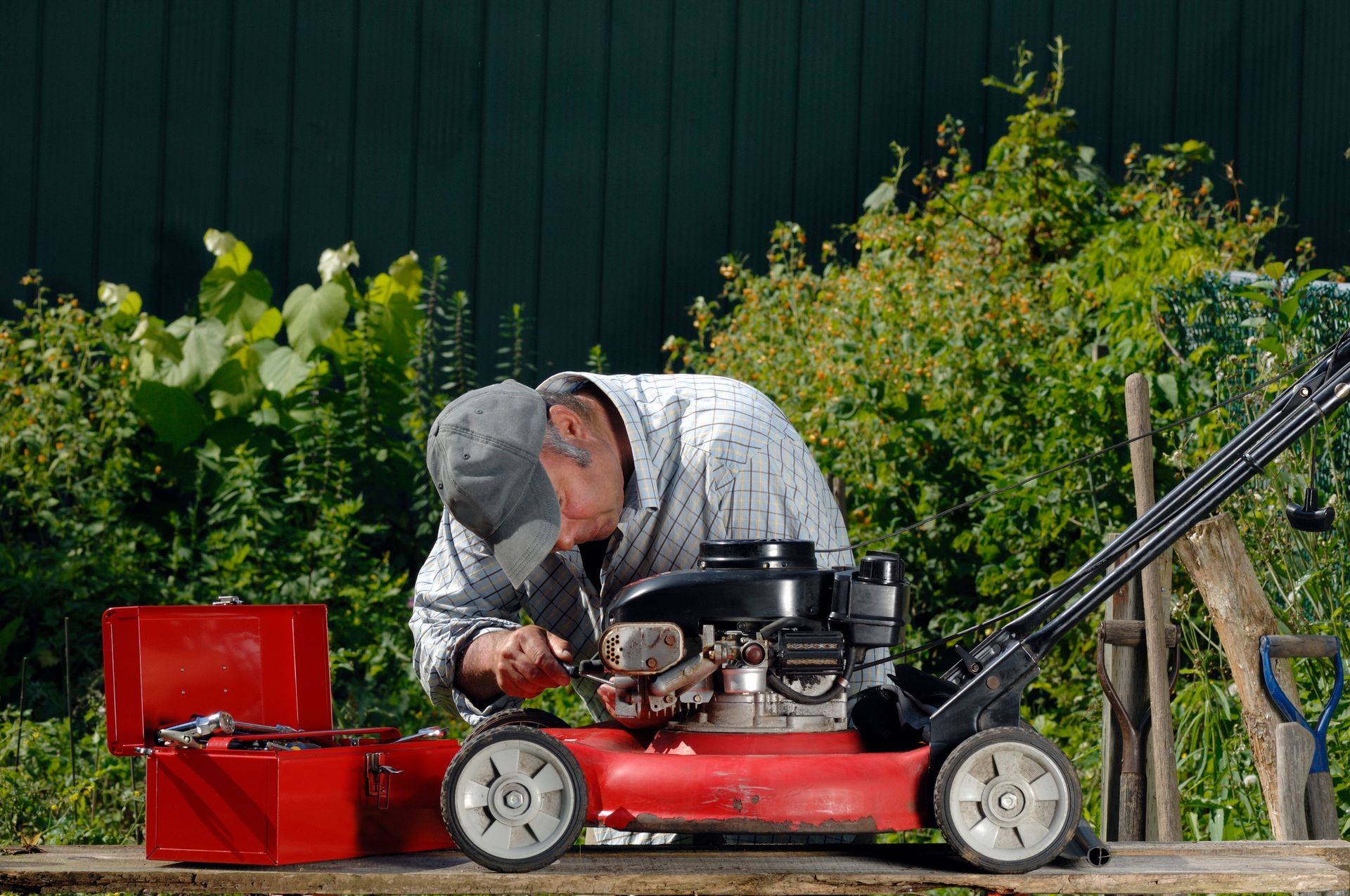 Man repairing a red lawnmower in a garden, with tools and greenery in the background.
