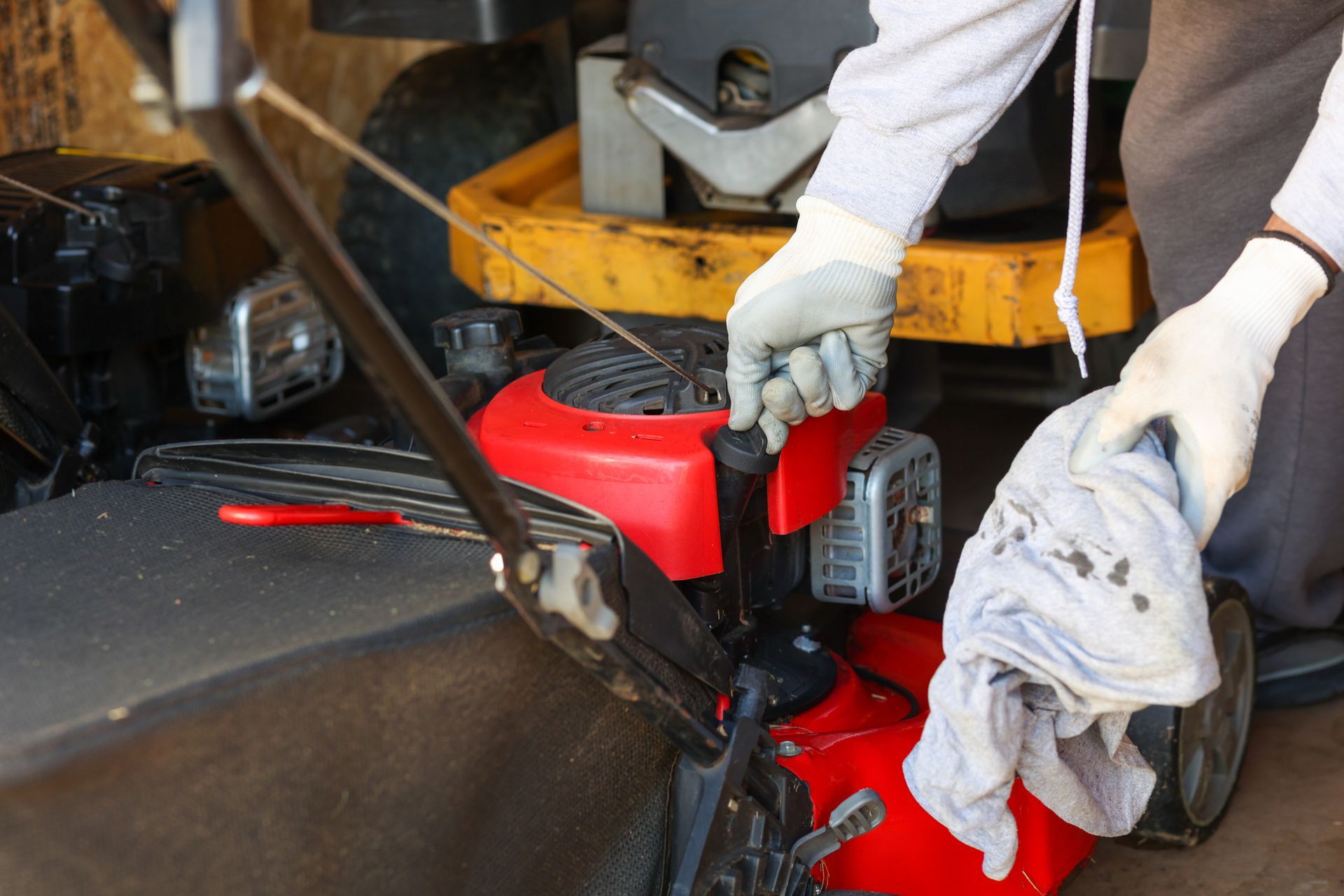 Person in gloves wiping a red lawnmower engine with a cloth, likely performing maintenance in an outdoor setting.