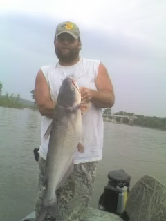 Person on a boat holding a large fish on a calm lake, with fishing gear nearby.