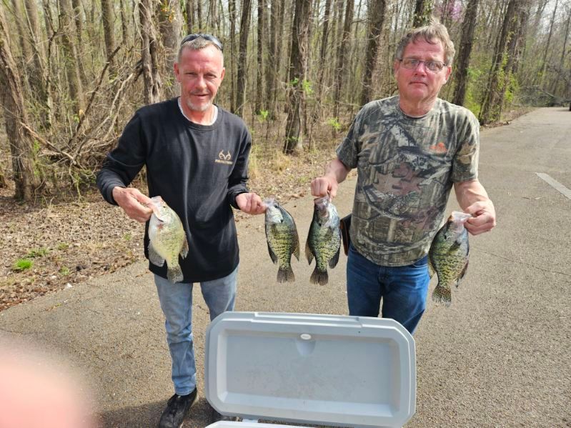 Two people hold several fish beside an open cooler on a wooded roadside.