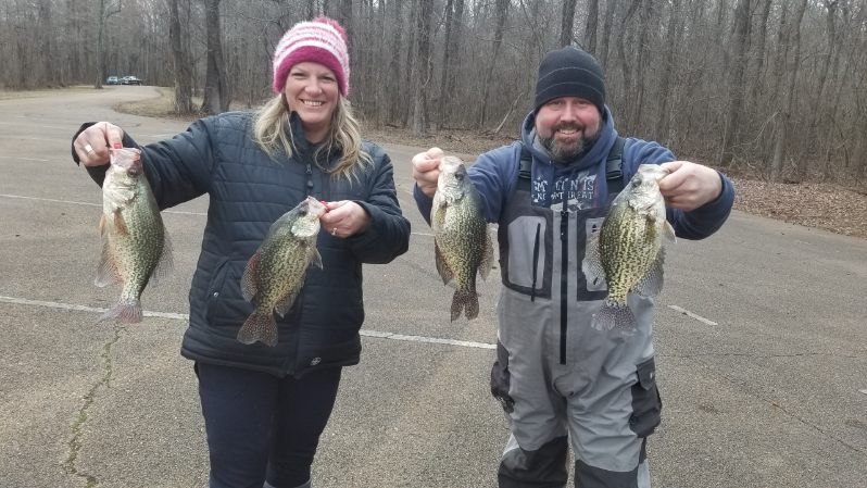 Two people in winter clothes holding up four fish outdoors on a paved area with bare trees behind them