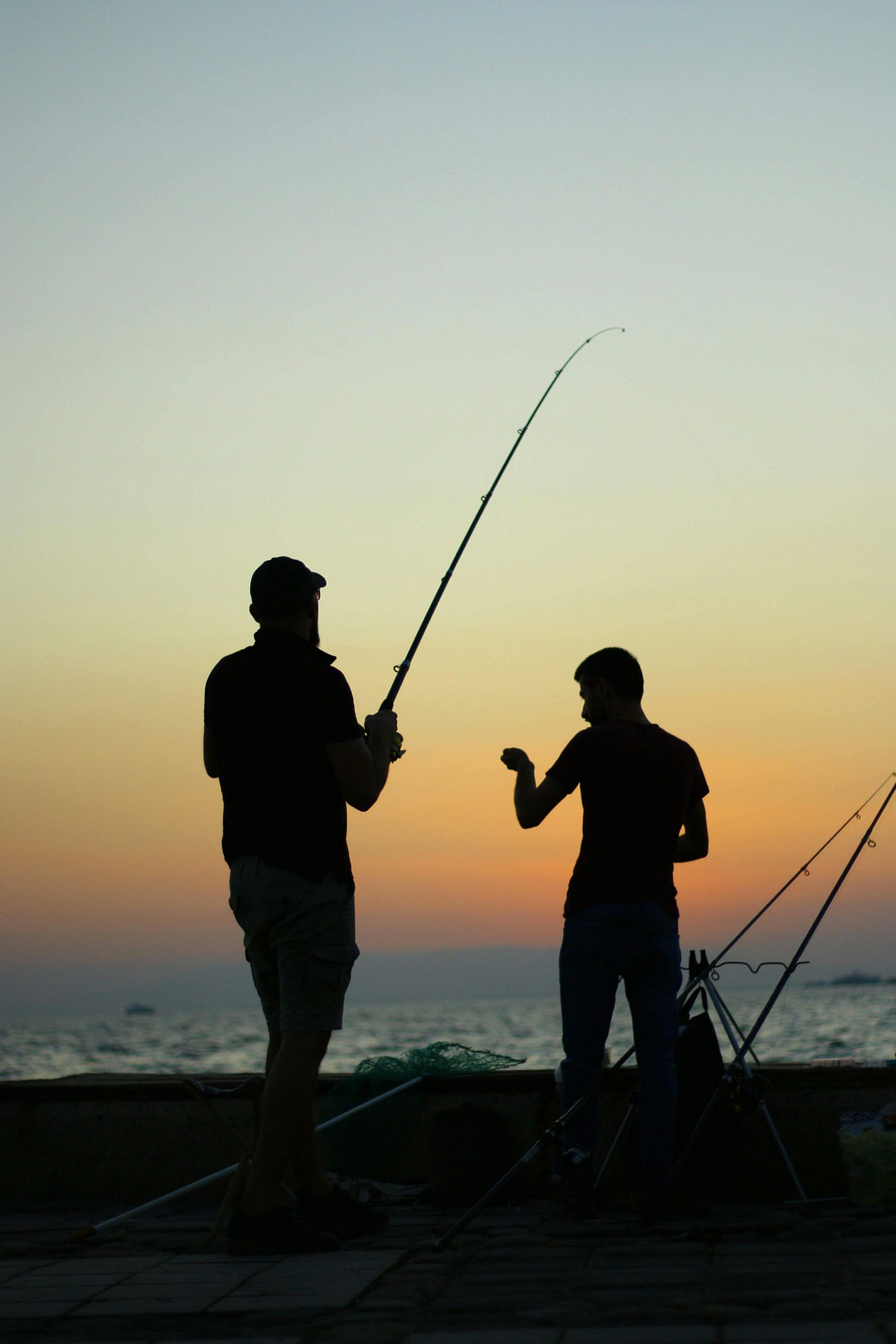 Two people fishing by the shore at sunset, silhouetted against an orange sky.