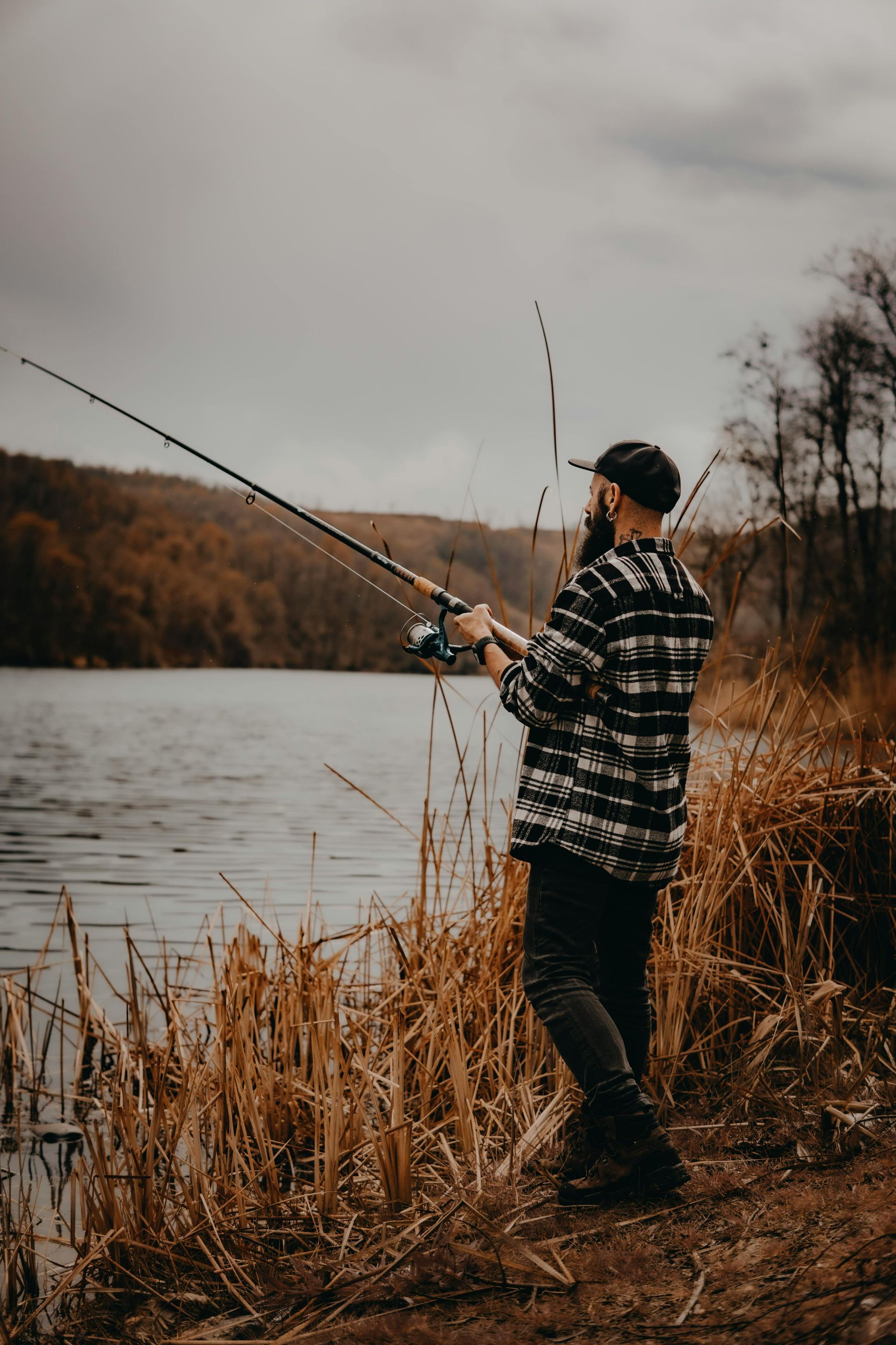 Person fishing on a lake shore in a plaid jacket under an overcast sky