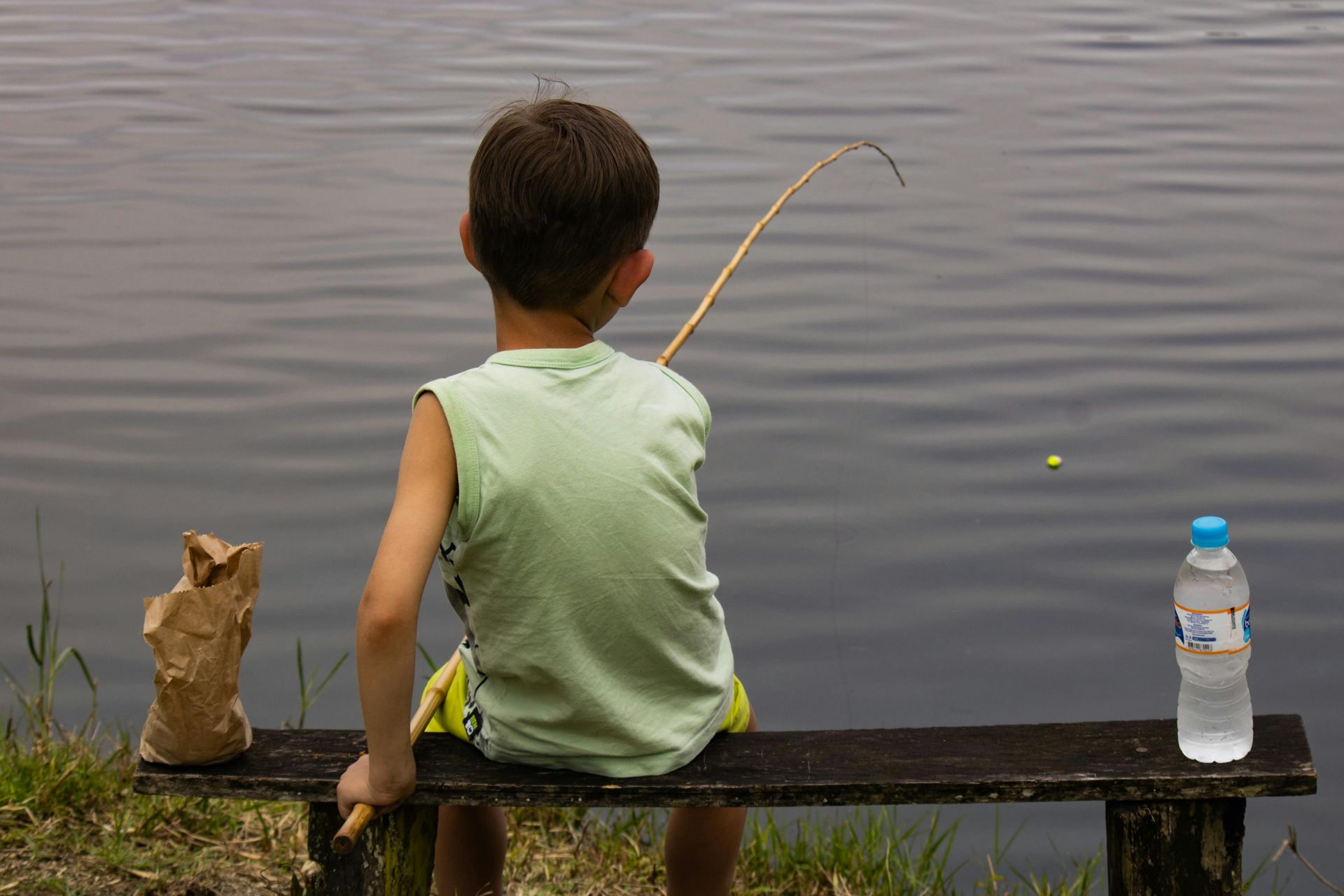 Child fishing from a bench by a calm lake, with a water bottle beside them.