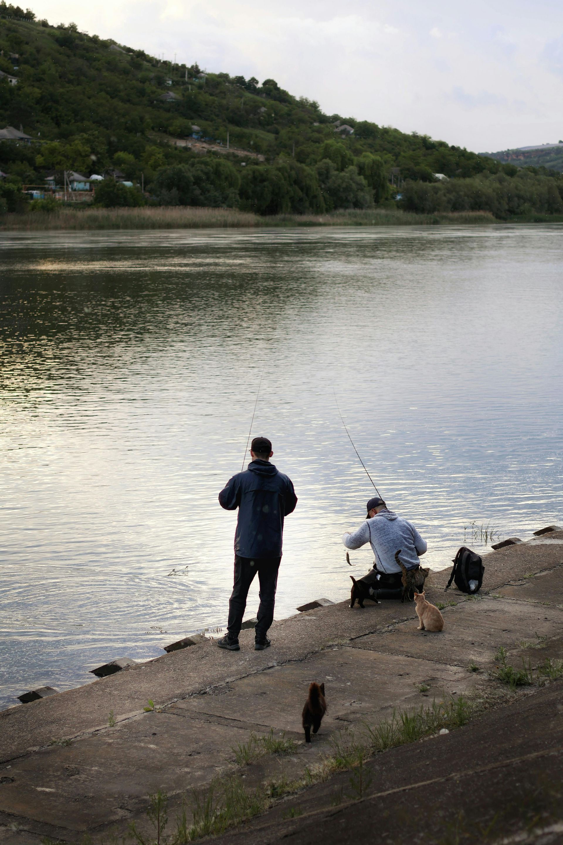 Two people fishing by a calm lake beside a wooded hillside