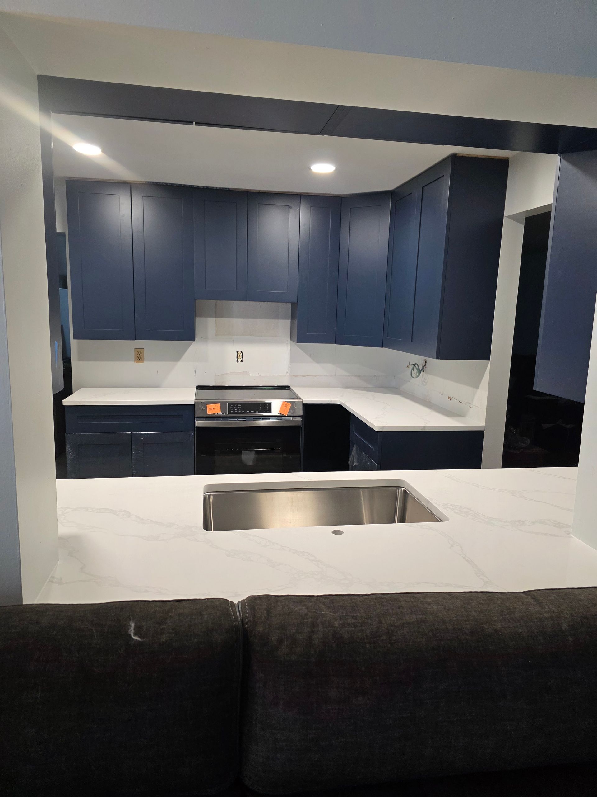 Blue kitchen cabinets with white countertops viewed from a room opening. Stainless steel sink.