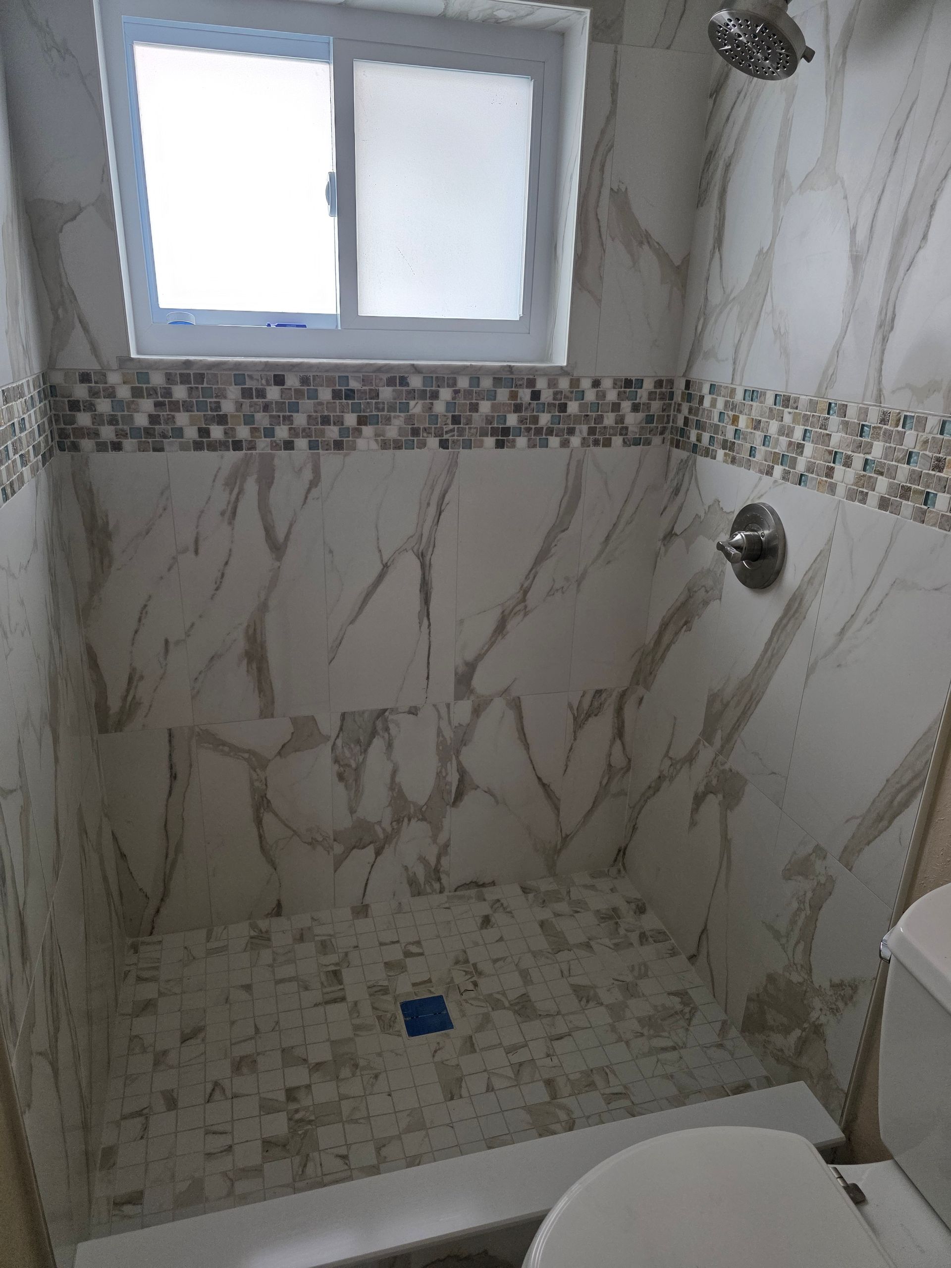 Shower stall with marble-patterned tiles, mosaic border, and window above. A chrome showerhead and faucet are installed.
