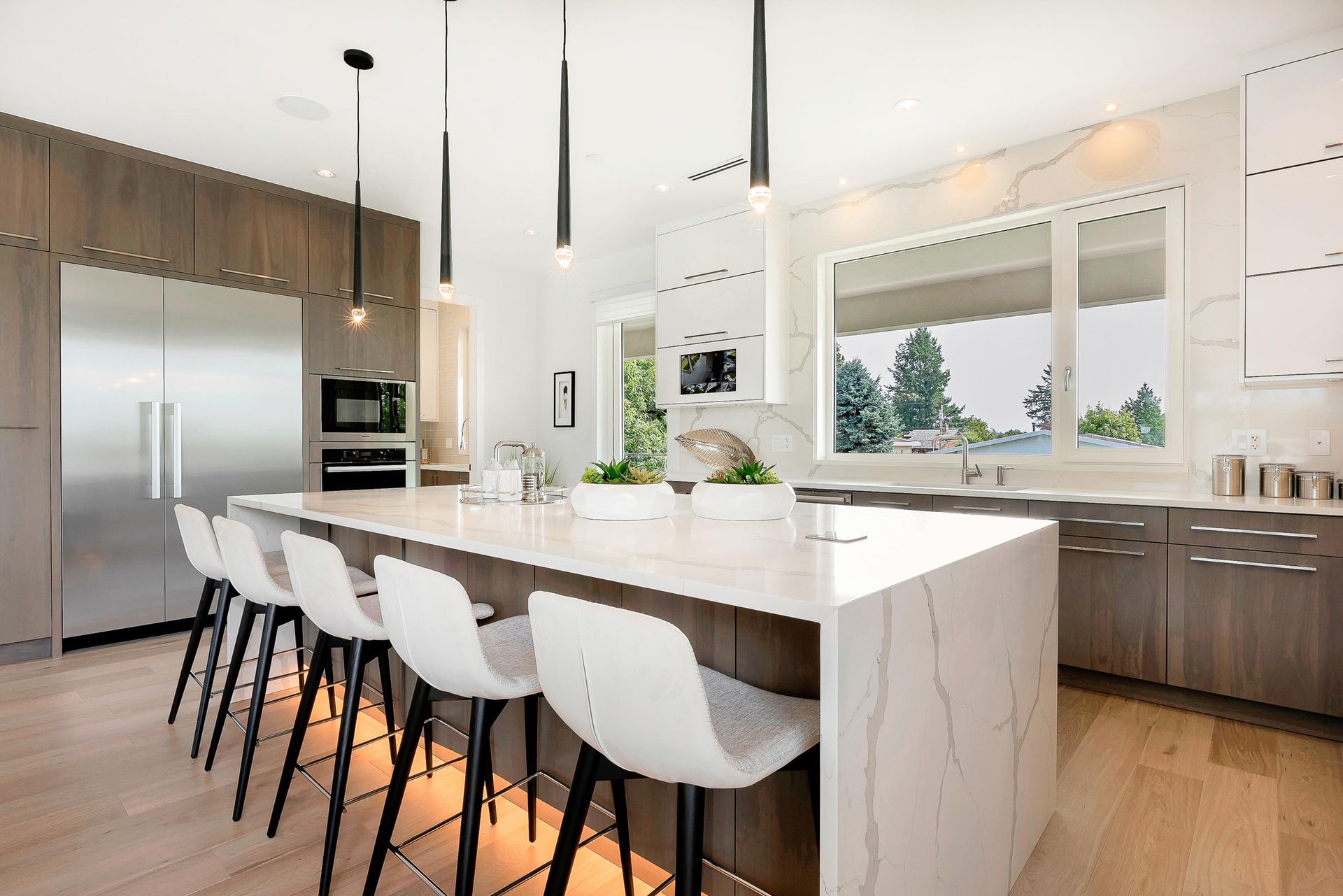 Modern kitchen with white island, black stools, stainless appliances, and large windows overlooking greenery