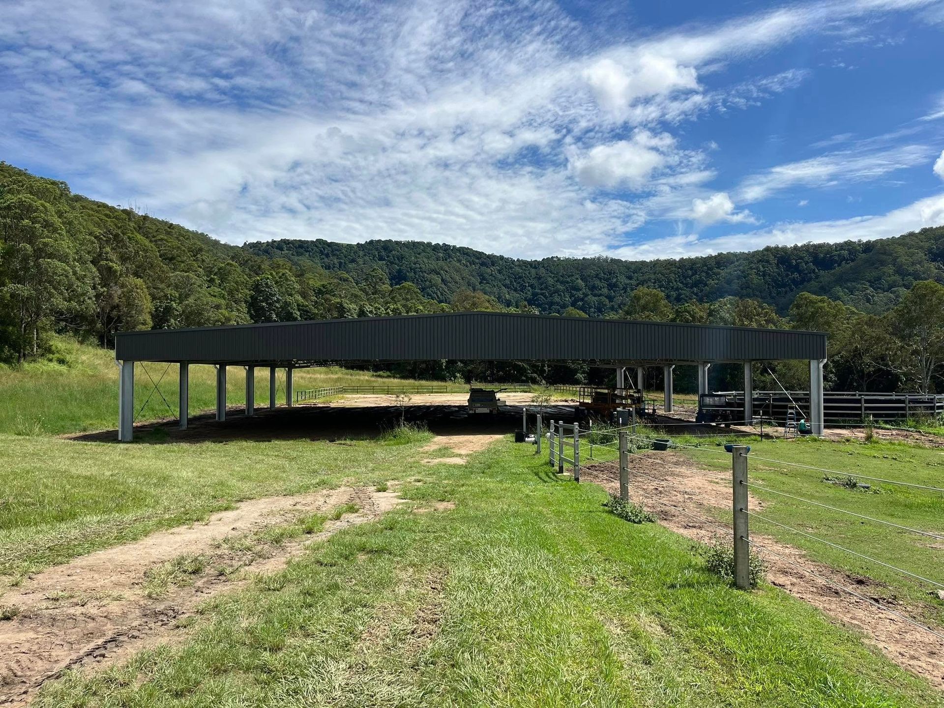 Open-sided metal shelter in a grassy field with a mountain backdrop under a cloudy sky. — Equine Stable Solutions In Diddillibah, QLD
