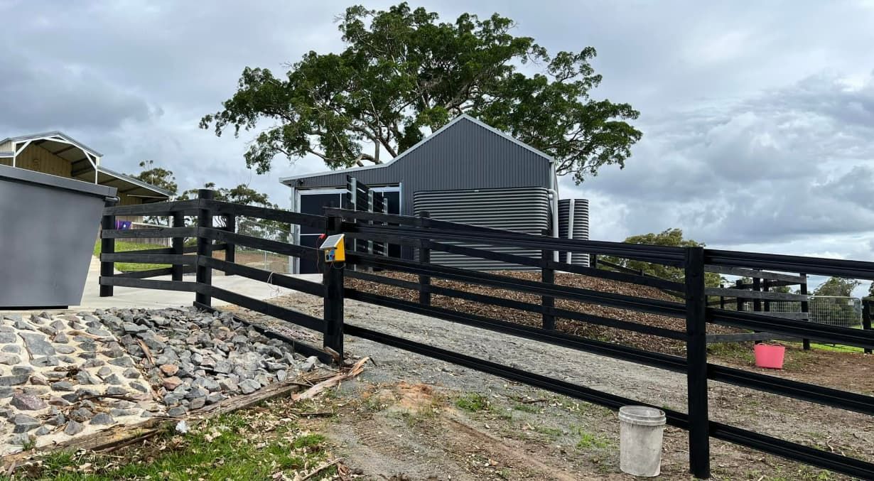 A Black Wooden Fence Surrounds A House On A Cloudy Day — Equine Stable Solutions In Brisbane, QLD