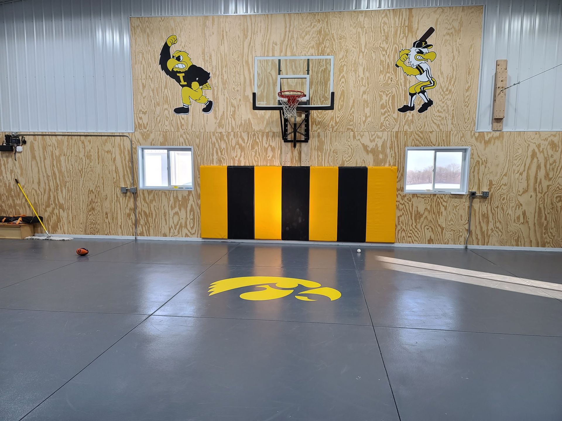 Empty indoor basketball court with yellow-and-black wall padding and a logo on the floor.