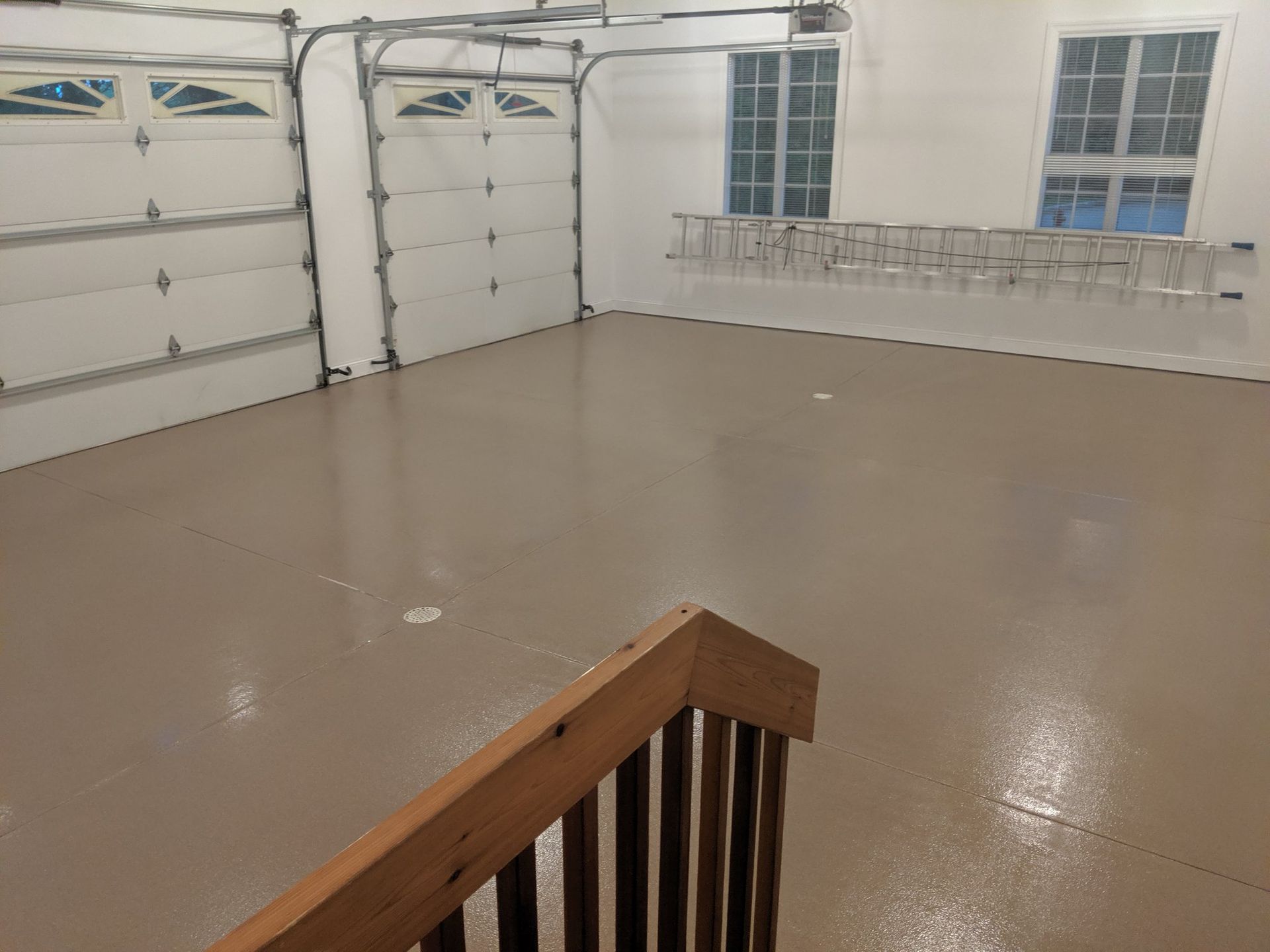 Empty garage with beige floor, white garage doors, windows, and a wooden stair railing in the foreground