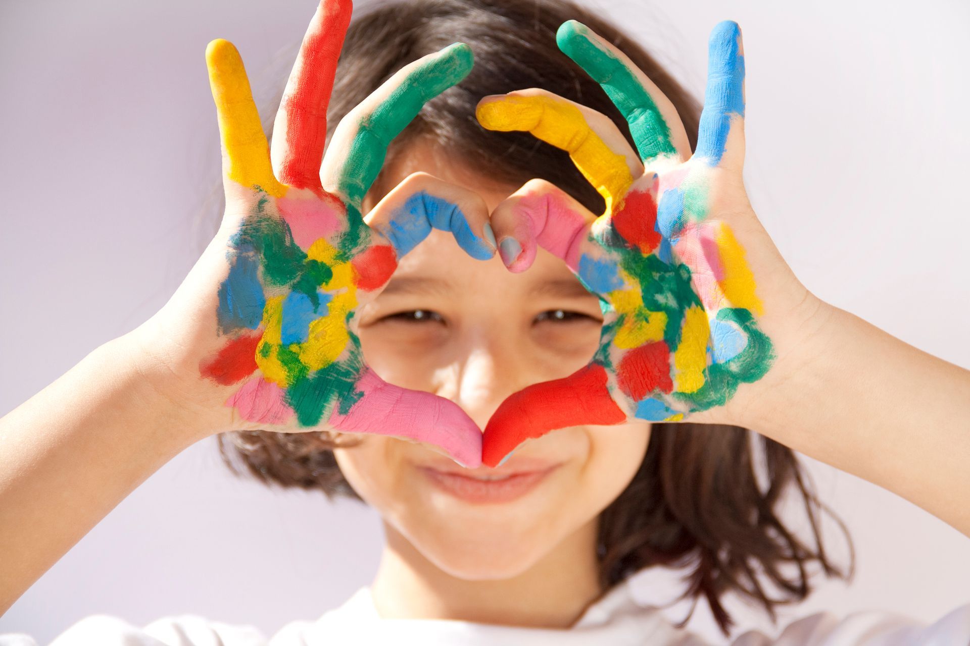 Girl smiles, making a heart with colorful, paint-covered hands.