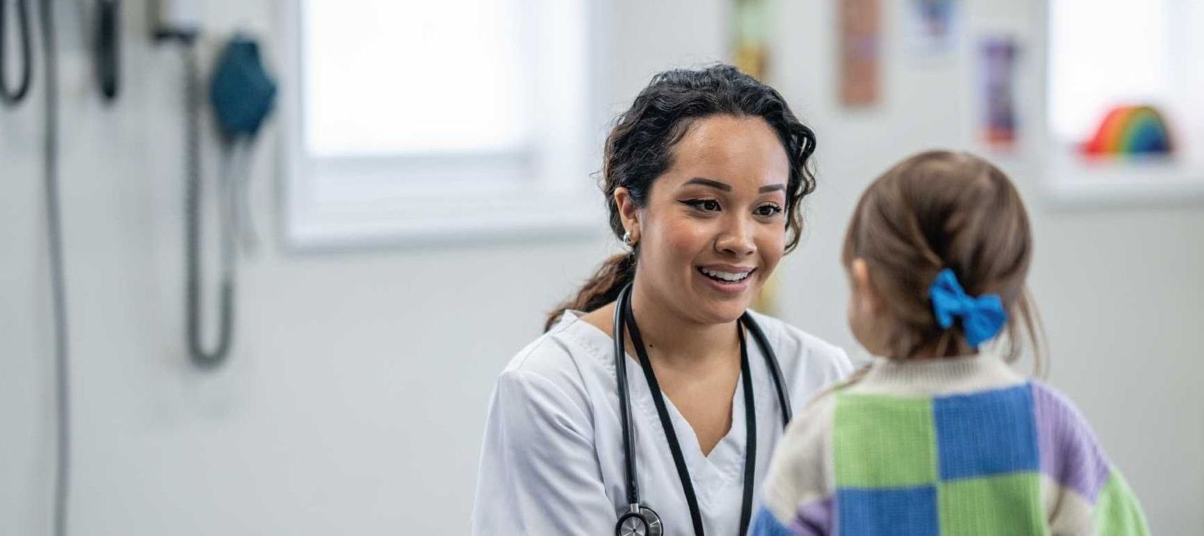 A doctor smiles at a young child in a light-filled clinic. The child wears a blue bow and a colorful sweater.
