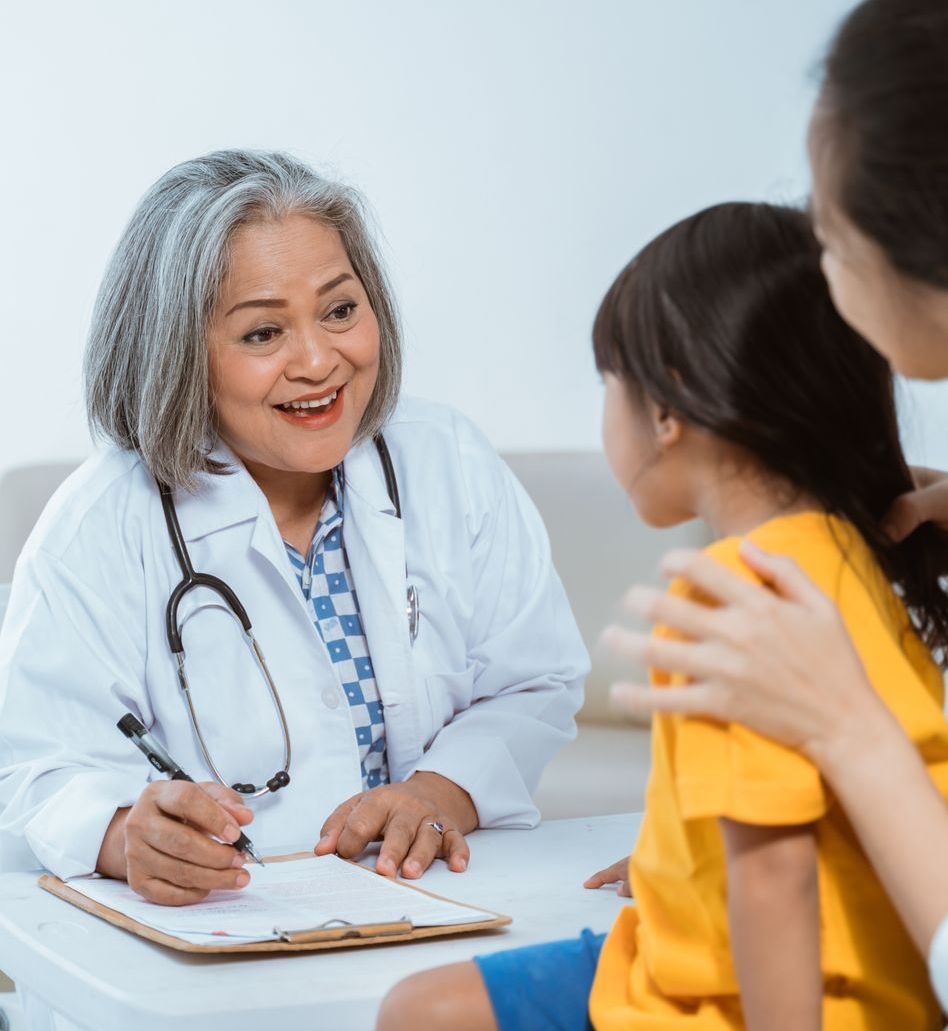 Doctor speaking to a young girl in a yellow shirt, mother present. They're indoors.