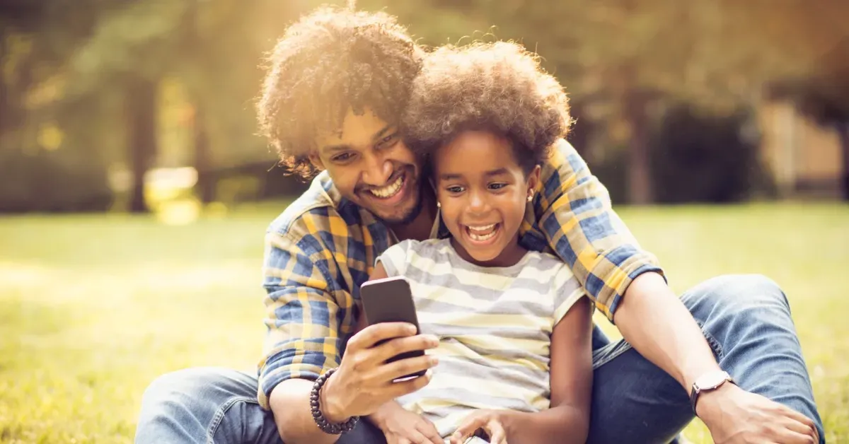 A smiling father and daughter with afros sit in a park, looking at a phone together.