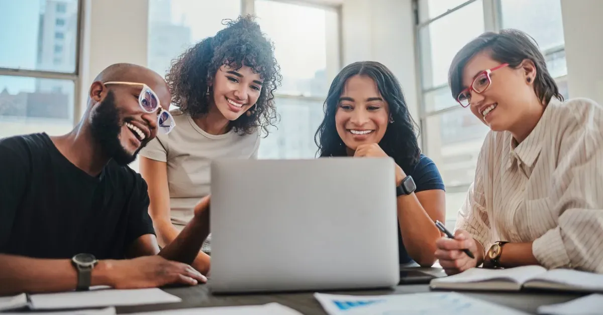 Four diverse coworkers smile while looking at a laptop together in an office.