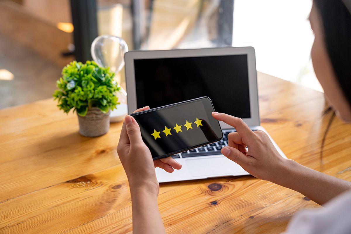 Woman holding phone with five-star rating in front of laptop on a wooden table.
