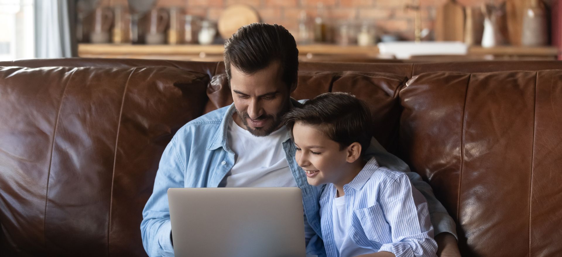 Father and son sit on a leather couch, looking at a laptop together.