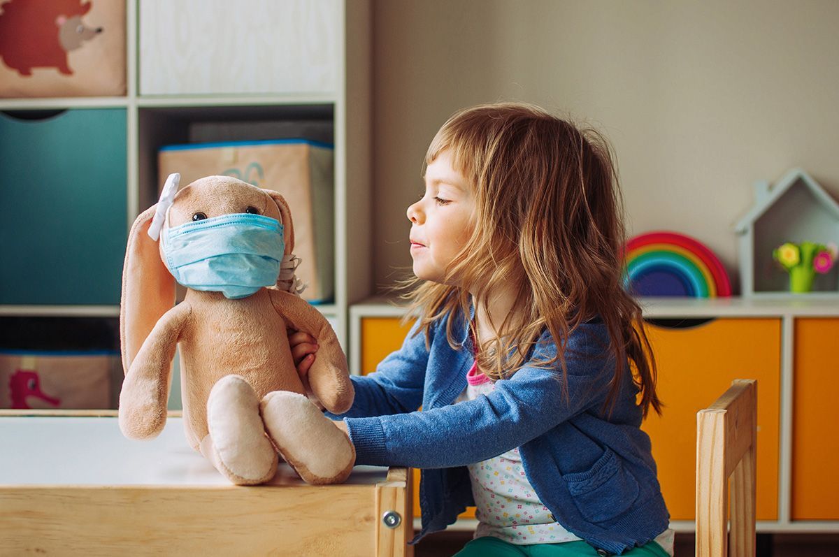 Girl placing a face mask on a stuffed bunny indoors with colorful decor.