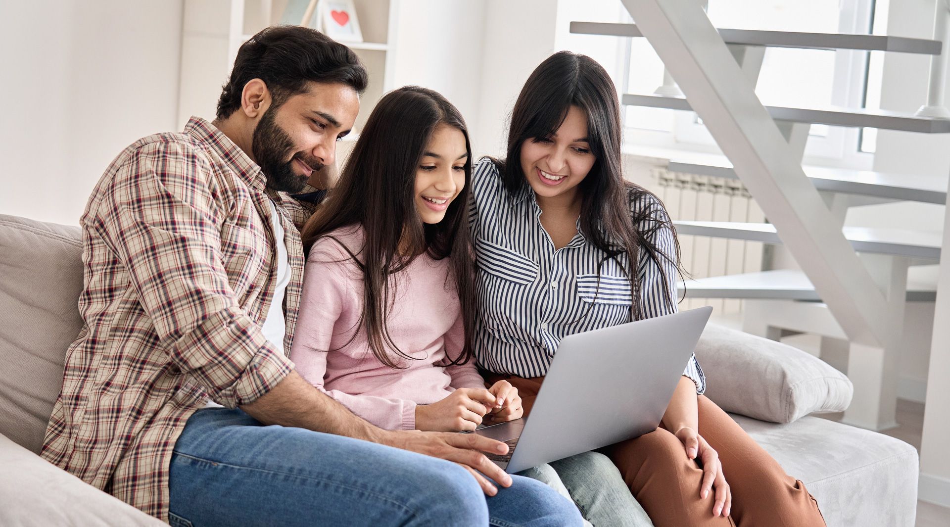A family sits on a couch together, smiling while looking at a laptop computer.
