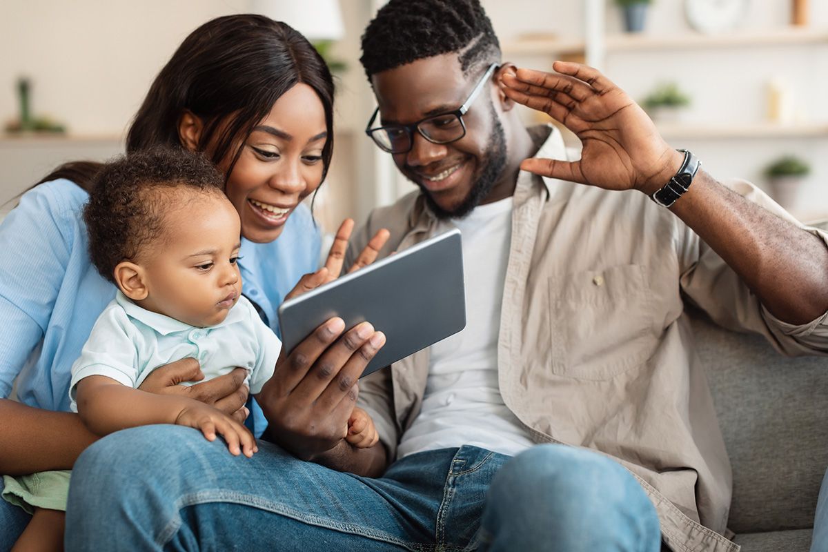 Family of three on a couch, looking at a tablet. Smiling parents, baby in arms.