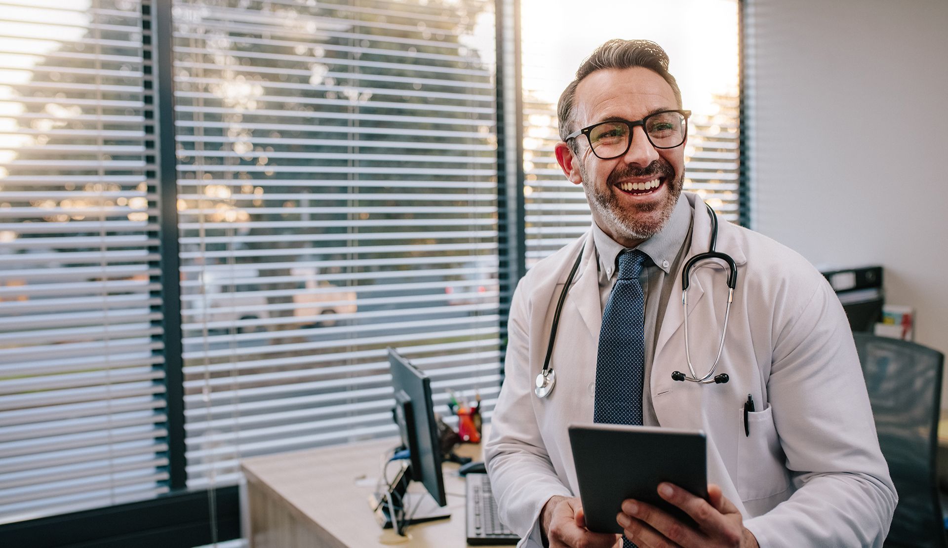 Smiling doctor holding a tablet in his office, wearing glasses and a stethoscope.