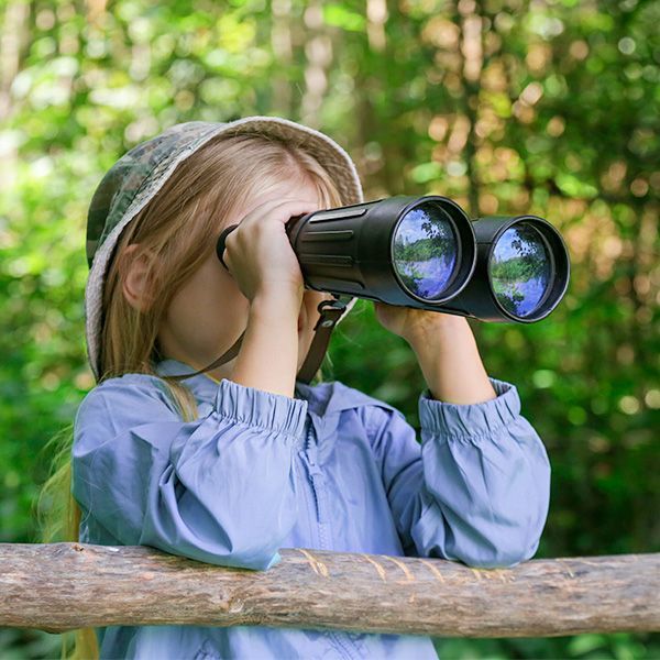 A young child, wearing a hat and blue shirt, looks through binoculars at a forest scene.