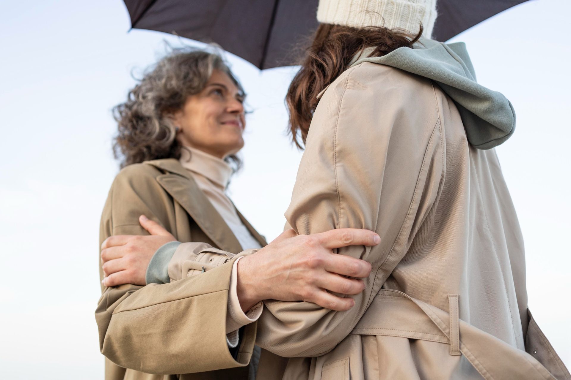 Two people embracing under an umbrella, wearing beige coats outdoors