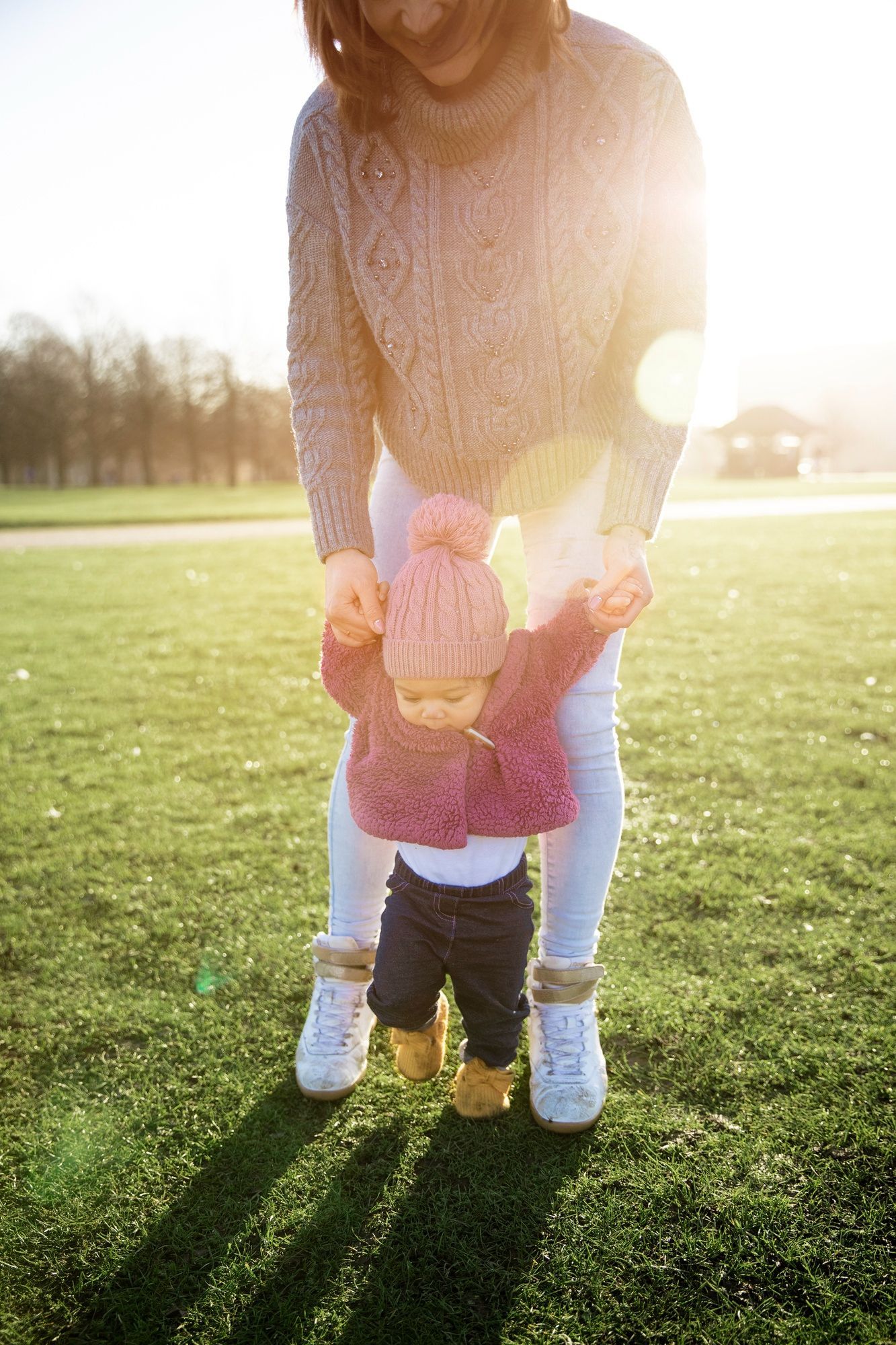 Adult holding a toddler’s hands while walking on a sunlit grassy field at sunset