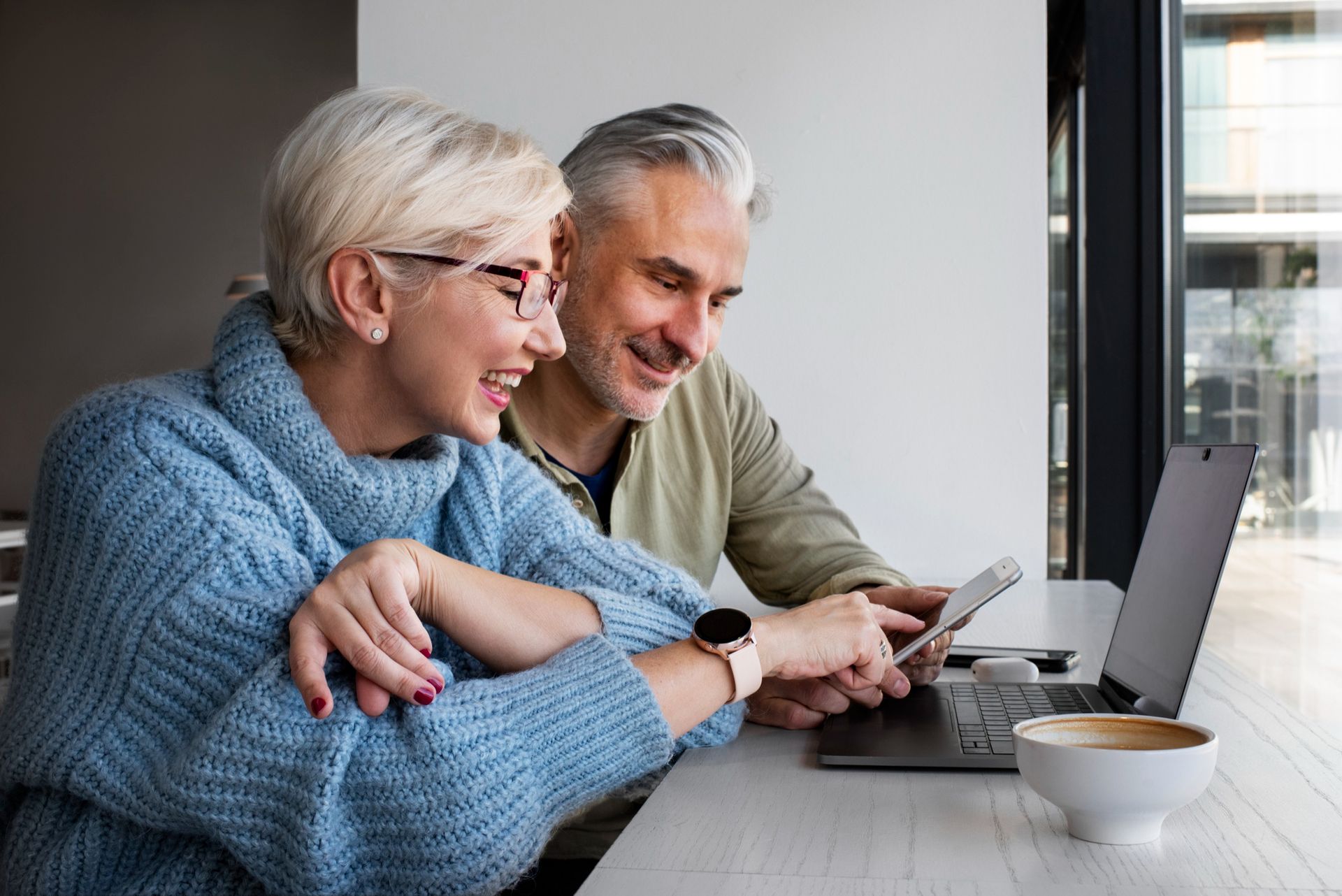 Two people smiling at a laptop while using a tablet in a bright café by a window.
