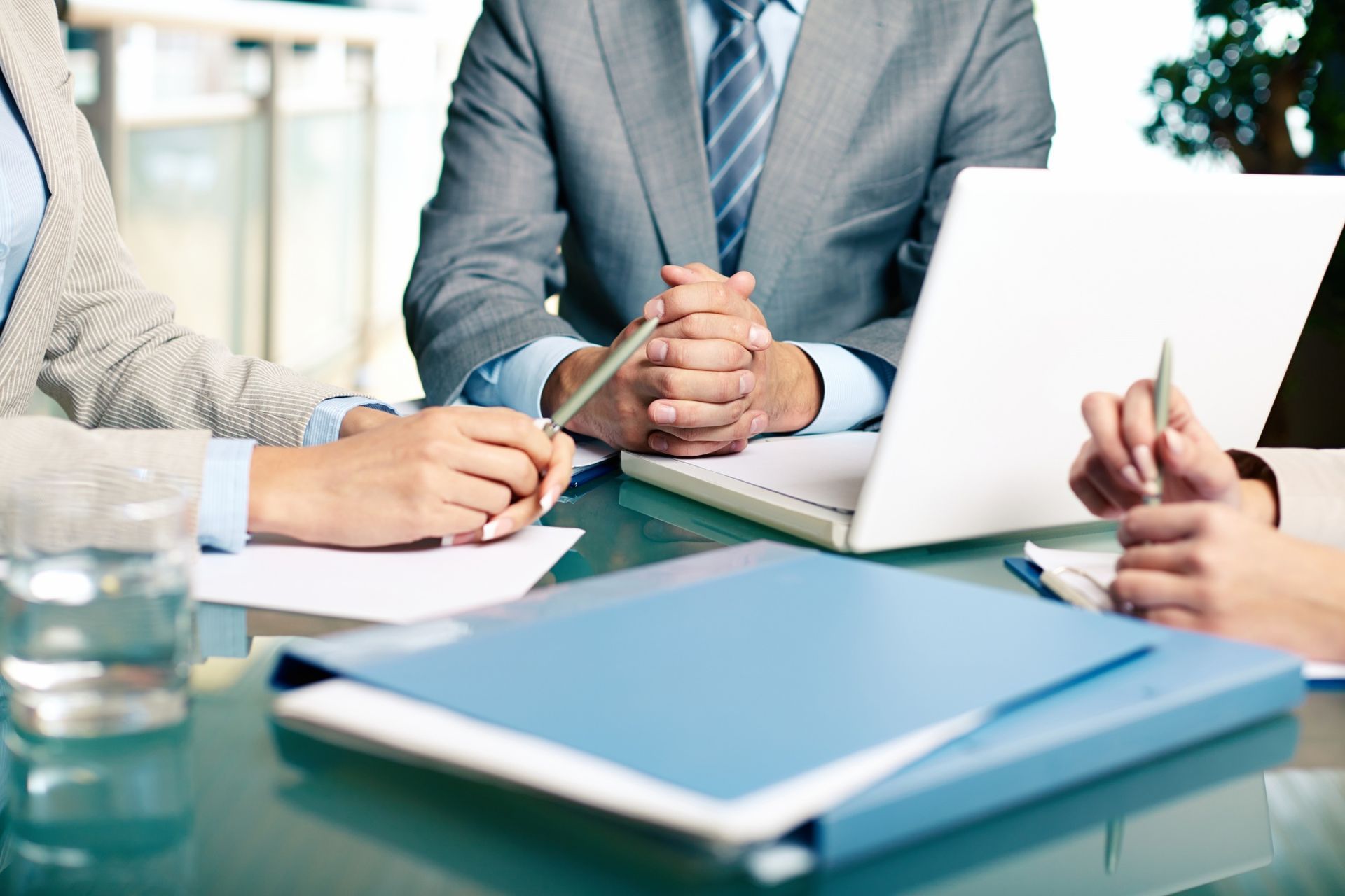Business meeting with three people around a table, laptops, notebooks, and documents in a bright office.