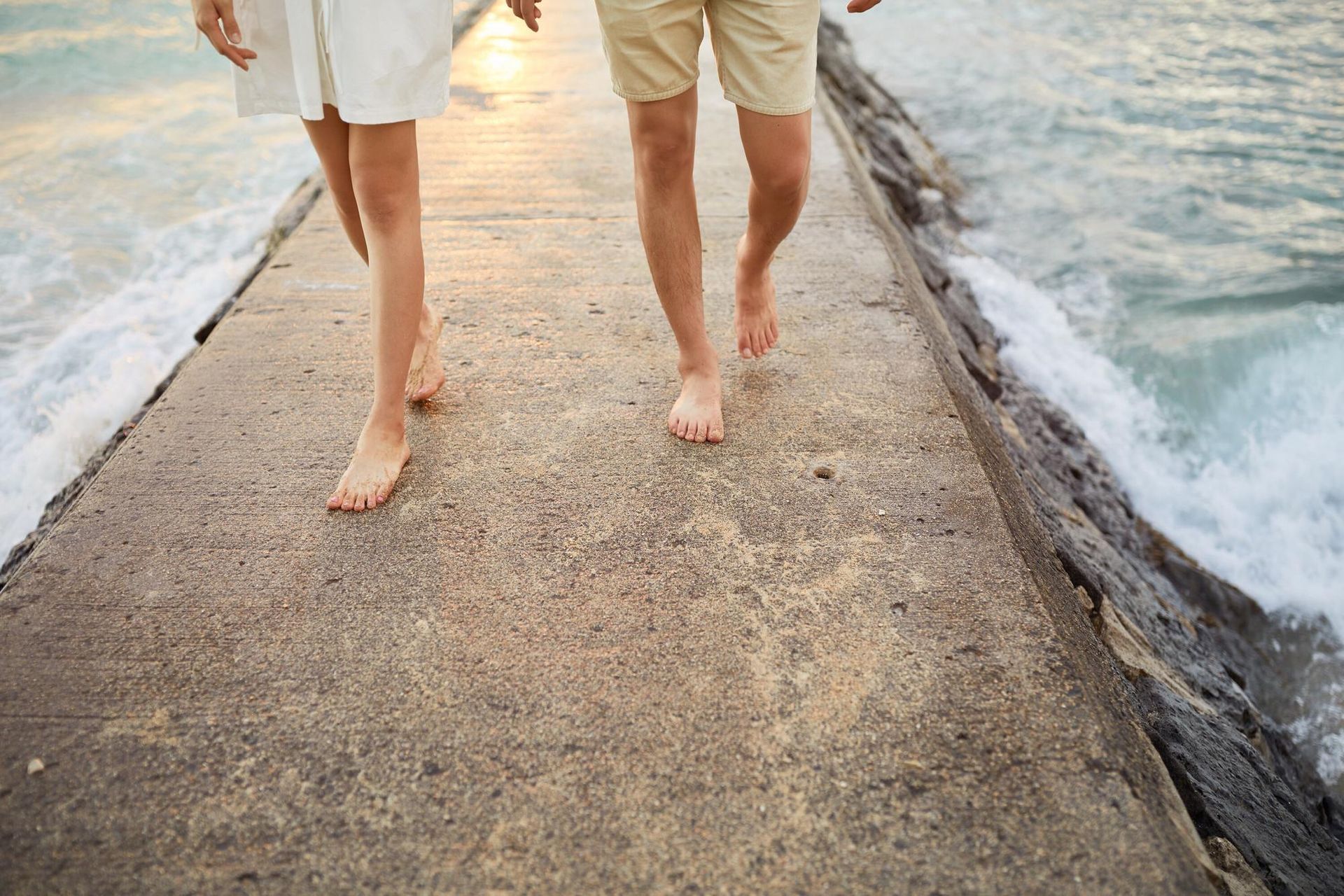 Two people walking barefoot on a narrow concrete path between ocean waves and sandy shore