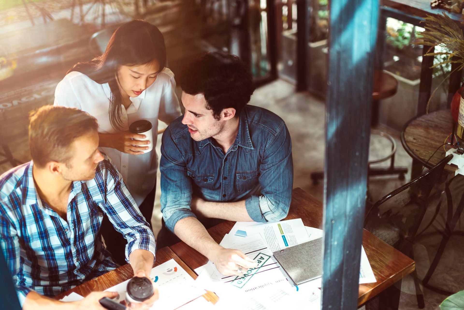 Three coworkers discuss papers and a laptop at a sunlit café table
