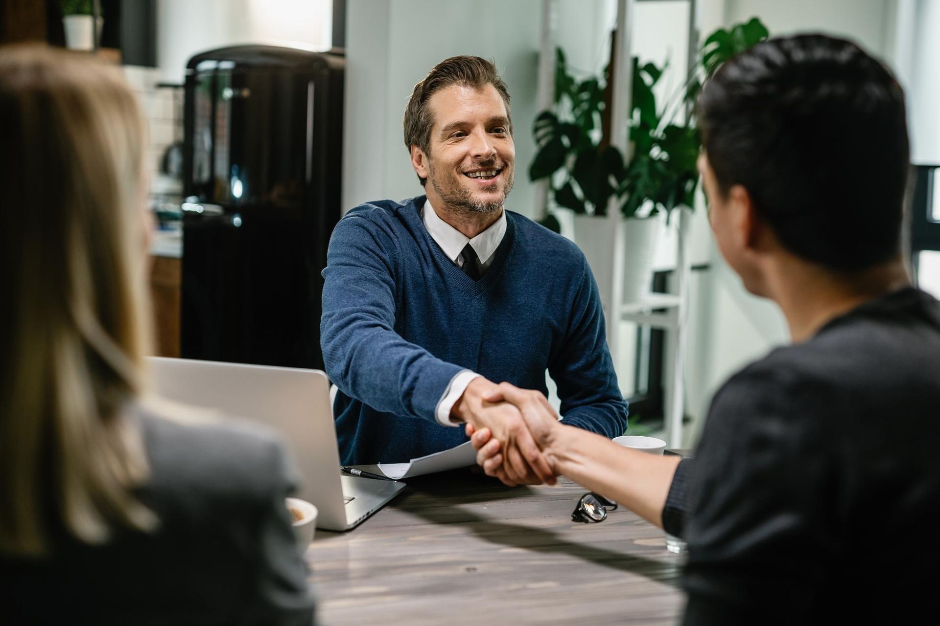 Business handshake across a desk in an office meeting with a laptop and plants in the background