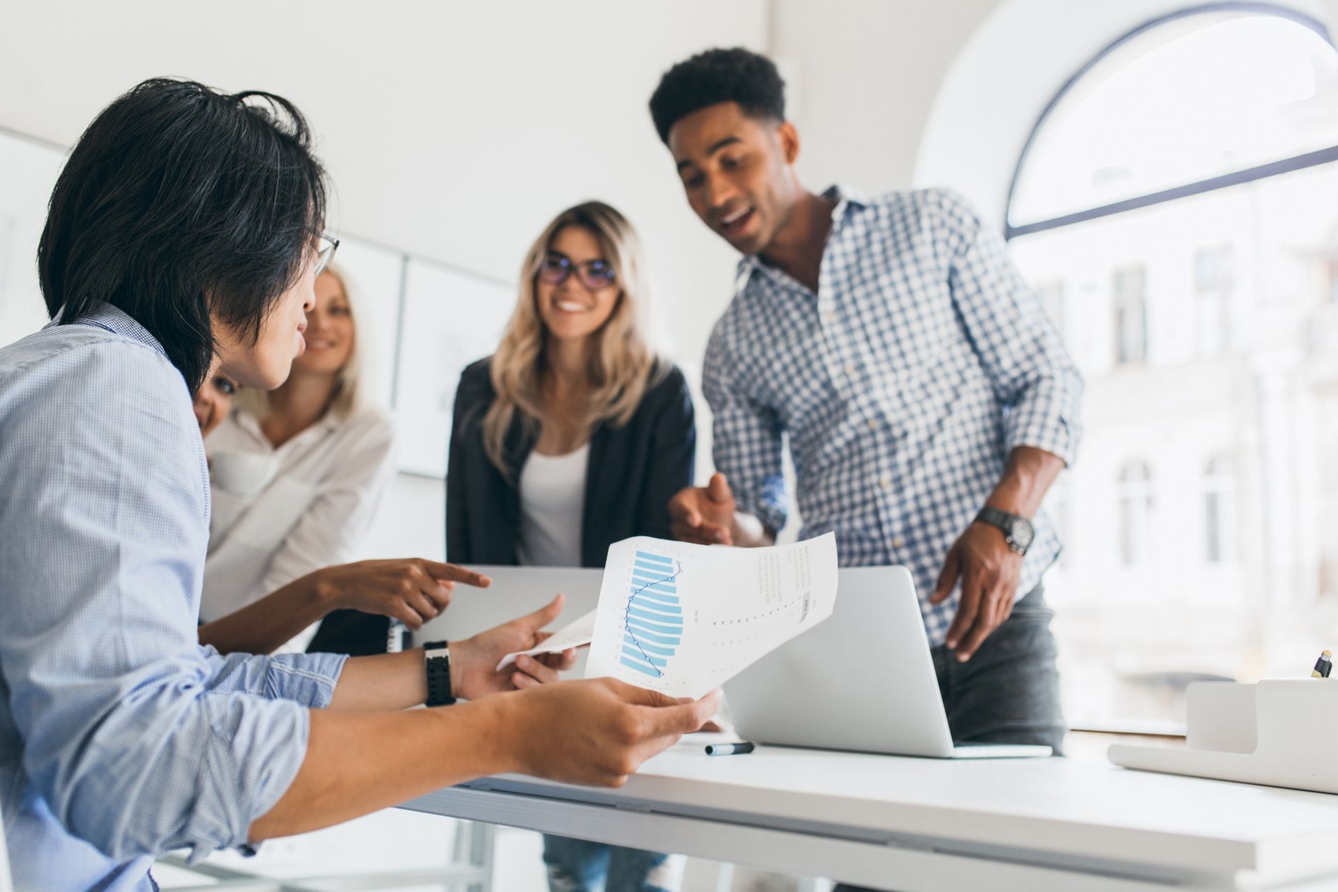 Coworkers meeting around a laptop in a bright office, reviewing charts and documents