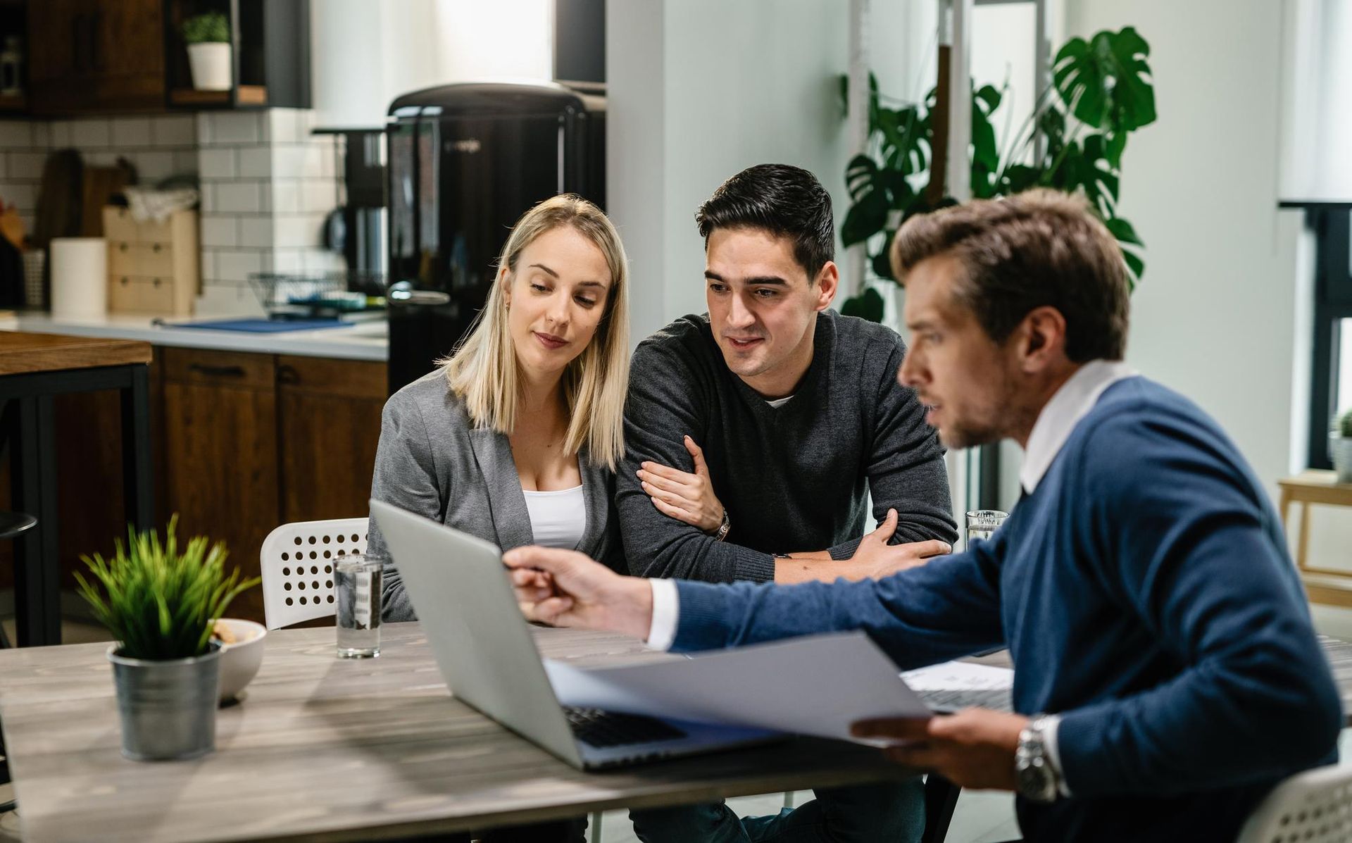 Three coworkers review a laptop at a table in a modern office kitchen