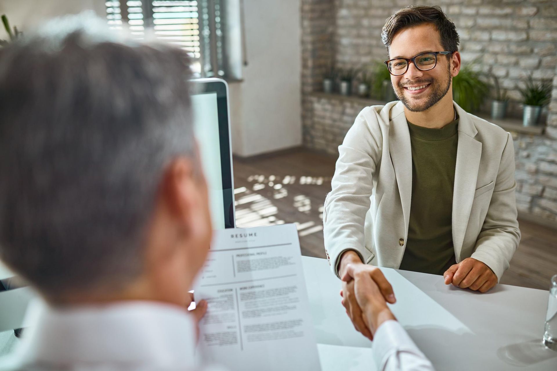 Two people shaking hands across a desk in a bright office interview setting