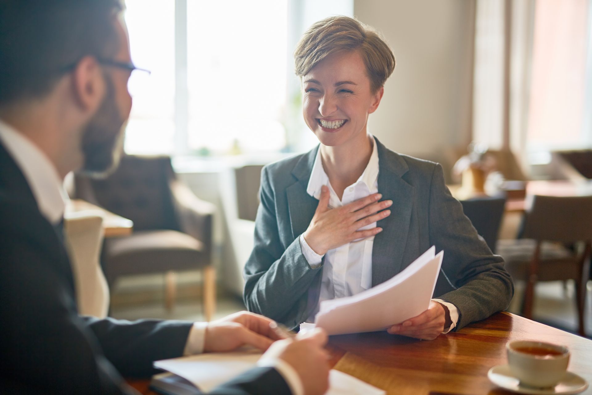 Two business people smiling and talking at a café table with documents and coffee cups