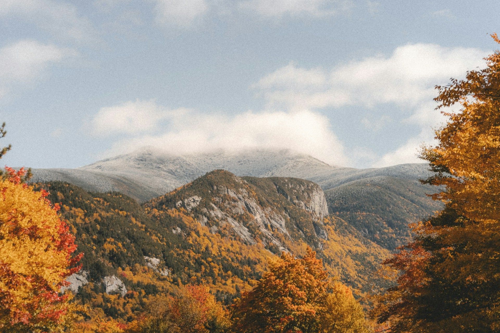 Autumn mountains under cloudy sky, with orange trees framing a rocky peak.
