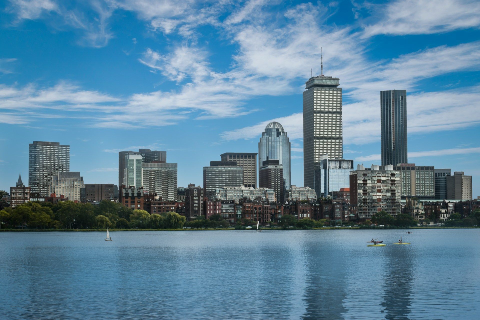 City skyline across a calm river under a cloudy blue sky, with several modern buildings and a bridge in the distance.