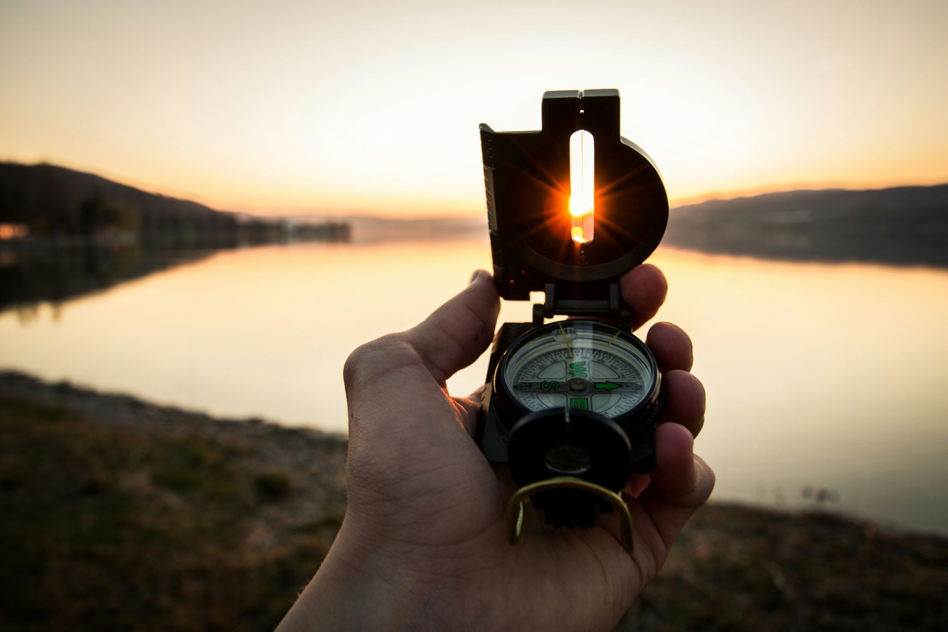 Hand holding a compass with the sun shining through it at sunset by a calm lake