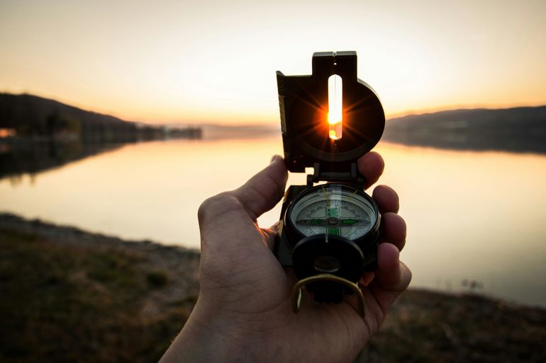 Hand holding a compass with the sun shining through it at sunset by a calm lake